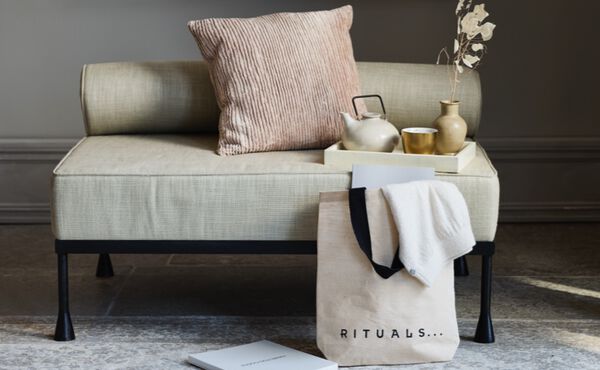 Reusable Rituals shopping bag with towel and notebook beside a sofa with a pink cushion and golden tray.